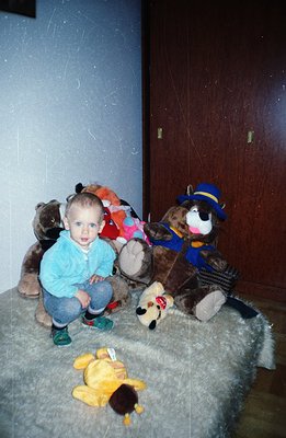 A young boy sits surrounded by stuffed animals on what appears to be a faux fur rug. He wears blue jeans and a light blue swe...