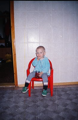 A young child sits in a red plastic chair, covered in red, likely fruit, residue. The child wears blue overalls, striped sock...