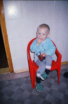 A young child sits in a red plastic chair, intently examining a jar of berries. Visible are green sneakers, dark socks, and a...