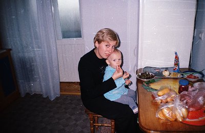A woman with short blonde hair holds a baby dressed in blue overalls. A richly laden table displays bread, fruit, cheese, and...