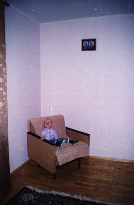 A young child sits in a mid-century modern armchair, captured in a domestic interior with textured wallpaper. The room featur...