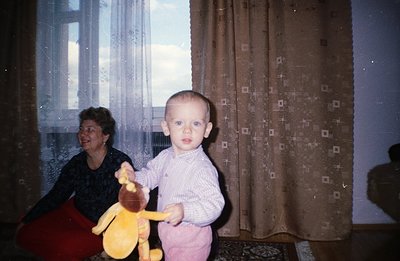 A young child, holding a plush deer, stands indoors with a woman seated in the background. Interior features include patterne...