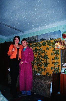 Two people stand close, one in a vibrant orange jacket and the other in a long purple robe with blue socks. A patterned wall ...