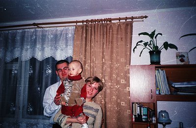 A family portrait: a man, woman, and young child stand near a window with patterned curtains. The child, dressed in red, is h...