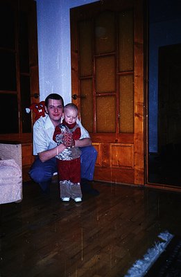 A man in a white shirt and jeans kneels, holding a baby in a patterned red and white outfit. A wooden door and parquet floori...