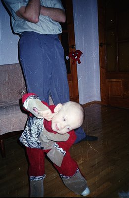 A toddler in red and patterned clothing appears to be playfully tipping forward, supported by the legs of an adult. Interior ...