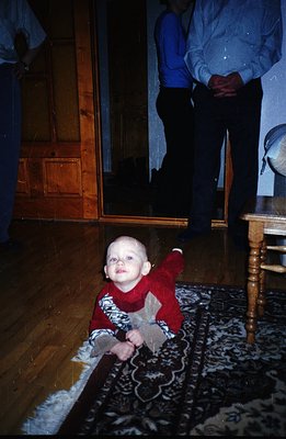 A baby in a red sweater crawls on a patterned rug within a domestic setting. A man in a blue shirt and dark pants stands near...