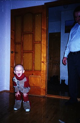 A young child with a distressed expression stands near an ornate, paneled interior door. The child is wearing a patterned jum...