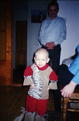 A young child, likely a toddler, stands in a warmly-lit interior, wearing a playful animal-print outfit. A man in a button-do...