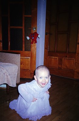 A young child with a shaved head kneels in a warmly-lit room, wearing a light purple, ruffled dress. A plush toy hangs on a d...