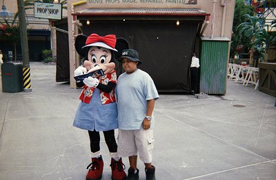 A young boy stands next to a costumed character resembling Minnie Mouse, posing for a photo. The backdrop suggests a themed a...
