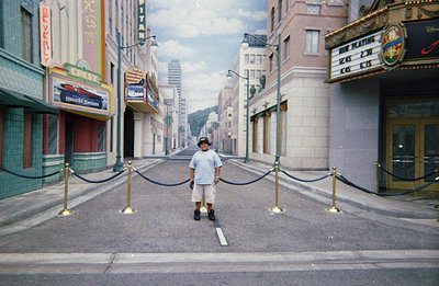 A young boy stands in the middle of a staged street, reminiscent of 1960s Hollywood. The backdrop features painted buildings ...