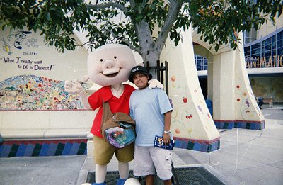 A young man in a baseball cap poses with a Porky Pig mascot. The backdrop features vibrant murals with script reading "What I...