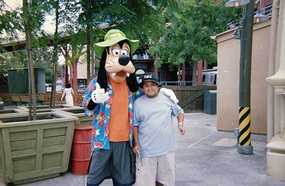 A man poses with Goofy, the Disney mascot, in an outdoor park setting, likely Disneyland or Disney World. Goofy wears a Hawai...