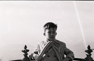 A young boy stands behind an ornate wrought-iron railing, gazing downward. He wears a double-breasted coat and scarf. The ima...