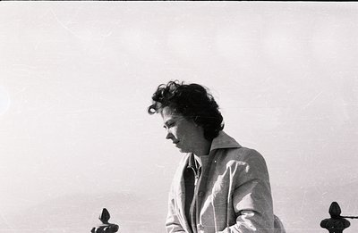 Man in a trench coat, captured in profile. Curly hair and a contemplative gaze characterize the subject; standing behind a wr...
