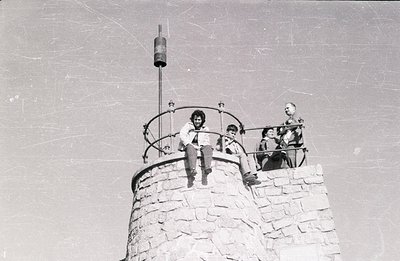 A group of four people sit atop a stone observation tower, likely in a mountainous region. The architecture features a circul...