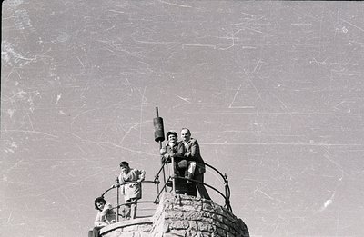 A grayscale photograph depicts three figures atop a stone structure with decorative wrought iron railings. Two adults, a man ...