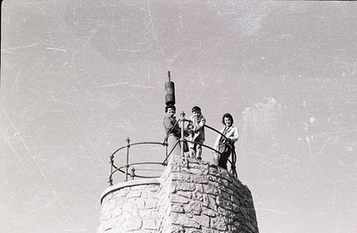 Three children pose atop a stone tower with a metal railing; one playfully balances a bucket on their head. Architecture sugg...