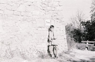 A young boy leans against a stone wall marked "Tubes," with a weathered wooden bench visible in the background. The monochrom...