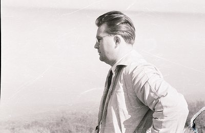 A man in a zipped jacket and tie observes a landscape. He wears glasses and has a neatly styled hairstyle. Appears to be a ca...