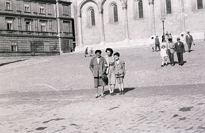 A black and white street scene captures three figures on a wide stone plaza. A woman and a boy, likely related, stand promine...