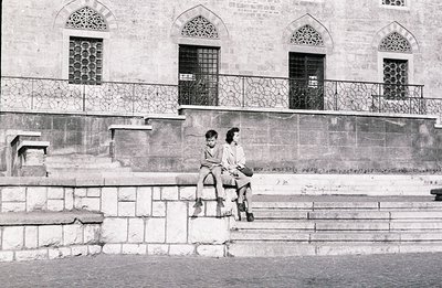Two children sit perched on a stone staircase leading up to a building with ornate arched windows and a decorative metal balc...