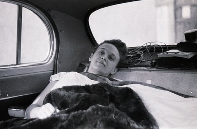 A young man rests inside a vehicle, likely a car trunk. He’s bundled in blankets and appears injured; his left arm is in a ca...