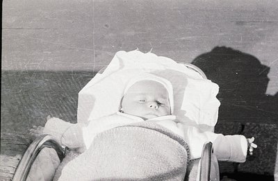 Infant asleep in a vintage pram, bundled in blankets & a close-fitting bonnet. A shadow is cast from the pram's frame. Likely...