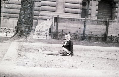 A young boy sits in a shallow depression on sandy ground, wearing a dark jacket and shorts. Ornate wrought-iron fencing and a...