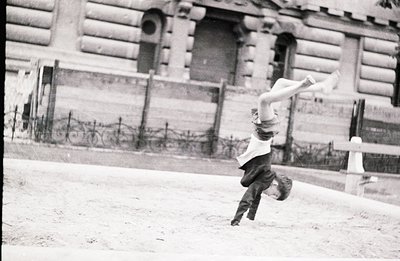 A young boy performs a handstand on a slightly sloped, gravel surface. He wears short pants and a button-down shirt. A detail...