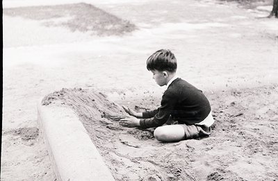 A young boy, dressed in a collared shirt and sweater, kneels within a shallow, concrete-lined depression, possibly a drainage...