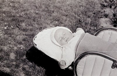 Infant lies peacefully in a vintage pram with striped lining. The baby wears a traditional white bonnet. Likely 1950s or 1960...