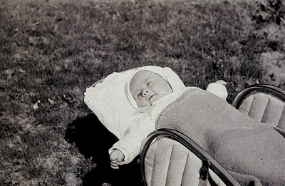 A baby, bundled in a white bonnet and blanket, rests in a vintage pram on a grassy lawn. The pram’s frame is visible, showcas...