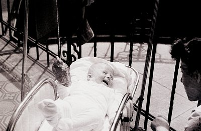 Infant in a stroller, likely outdoors, with patterned tile flooring visible. The child, dressed in white, appears to be laugh...