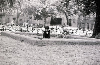 Two boys engaged in play within a walled park area. One kneels, examining the ground; the other sits, using a tool to manipul...