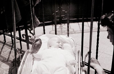 A baby lies in a vintage, ornate pram, eyes closed in apparent contentment. A woman’s hand and arm are visible, presumably pu...