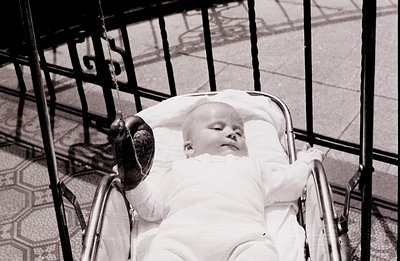 A serene infant, swaddled in white, sleeps peacefully within a vintage chrome and rattan pram. The stroller sits before a wro...