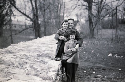 A young boy sits on a man’s shoulders atop a stone retaining wall, set against a backdrop of bare trees. Both are wearing mid...