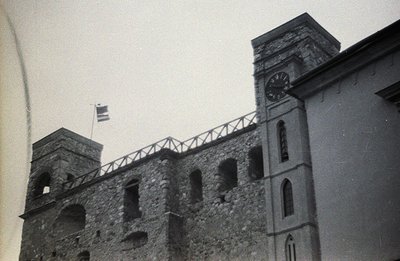 Monochrome photograph depicts a stone fortress or castle wall with a clock tower, likely European. Open crenellations line th...