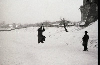 A man in a dark coat and hat leaps into a snowy landscape, seemingly propelled upwards. A second man stands observing, slight...