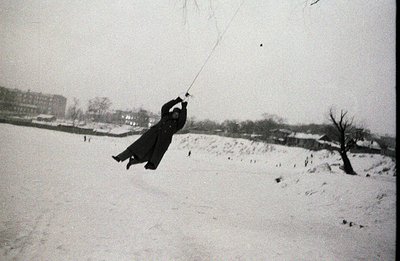 A person in a long coat swings from a rope across a snow-covered field. The pose suggests mid-swing, with blurred motion. Bac...