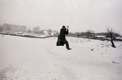 A person, bundled in a dark coat and hat, leaps mid-air while suspended from a rope. Snowy landscape with apartment buildings...