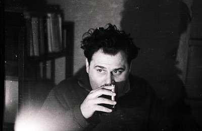Intimate portrait of a man with tousled hair, holding a drink and lit cigarette. Bookshelves fill the blurred background. Sta...