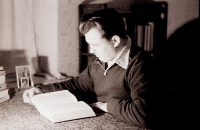 A young man, seen in profile, intensely reads a large, open book seated at a table. Shelves of books form a blurred backdrop....