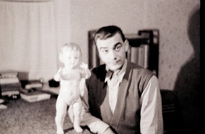 A man in a button-down shirt & vest poses with a small, plaster statuette of a child. The setting appears to be a room with b...