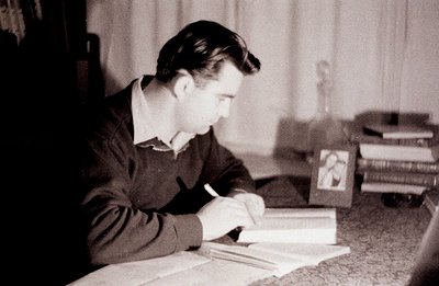A man in a dark sweater writes at a desk with a framed photograph and books visible. The composition features side profile, s...