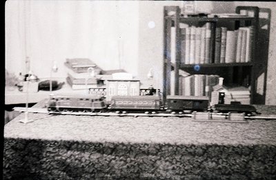 Detailed black and white photograph shows a miniature train set displayed on a table, with a large bookcase in the blurred ba...