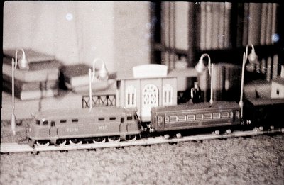 Detailed black and white photograph captures a miniature railway scene. A locomotive engine leads a passenger car on tracks, ...