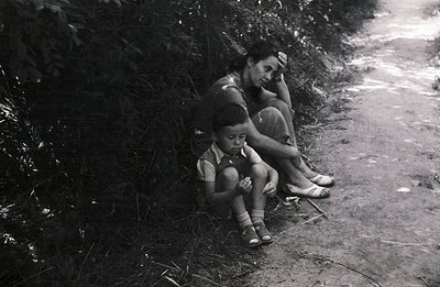 A mother and young boy sit nestled against foliage on a dirt path. The boy wears sandals and a collared shirt, while the moth...